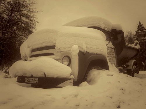 An old truck covered in snow, partially buried, with trees in the background and a vintage feel.
