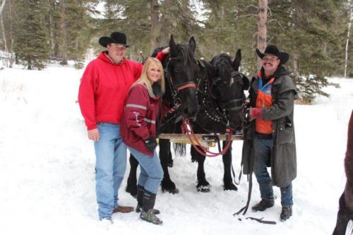 Three people in winter attire stand beside two horses in a snowy forest, with trees in the background.