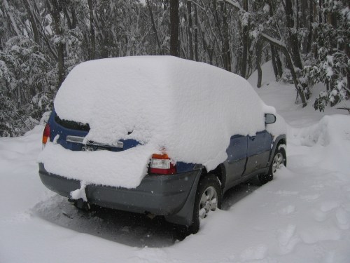 A car covered in thick snow, parked in a snowy forest with trees in the background.