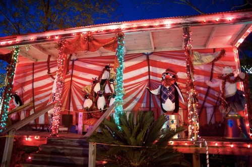 A festive scene featuring decorated penguin and polar bear figures on a porch, illuminated by colorful lights.