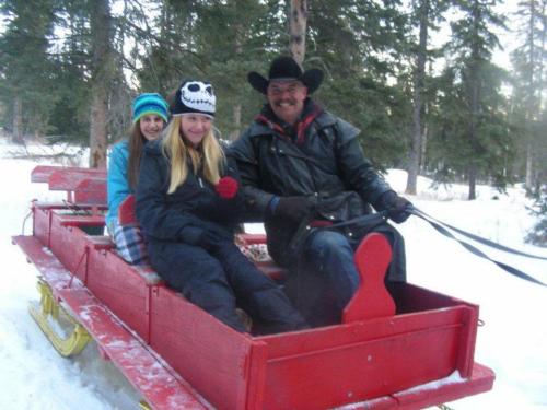 A man in a cowboy hat sits with two girls on a red sled in a snowy forest. Trees surround them.