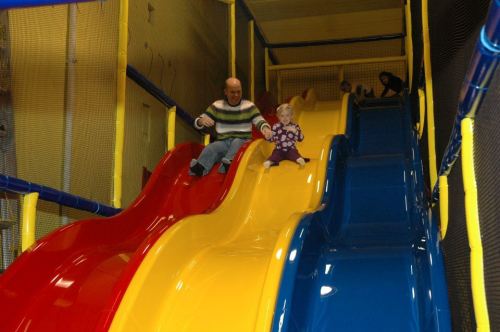 A child and an adult slide down colorful slides in a play area, with another child playing in the background.