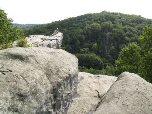 A person sits on a rocky ledge overlooking a lush green valley and distant hills under a bright sky.
