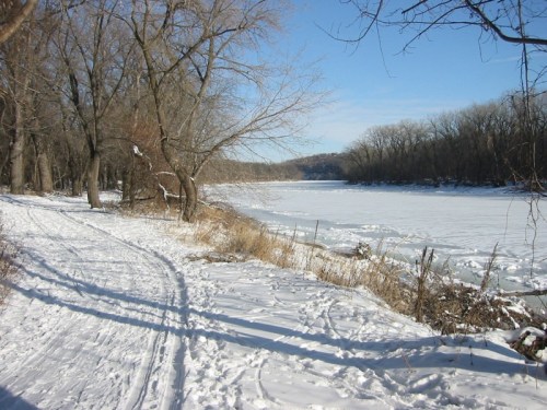 A snowy riverbank scene with bare trees and a frozen river under a clear blue sky.