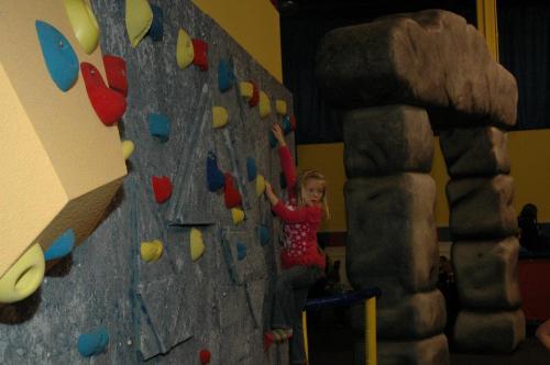 A young girl climbs a colorful indoor rock wall, with a large stone structure nearby.