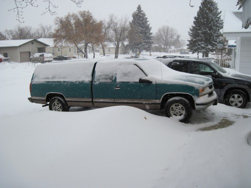 A teal truck covered in snow, parked in a driveway with snowdrifts and a black vehicle nearby. Snow is falling.