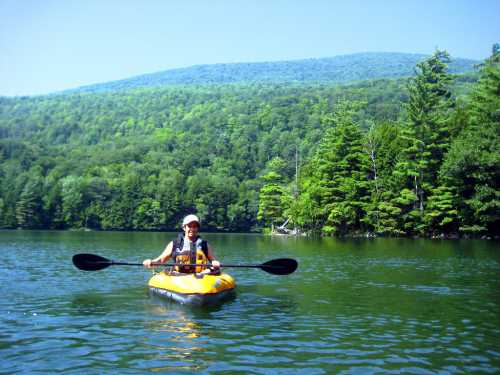 A person paddles a yellow kayak on a calm lake surrounded by lush green trees and mountains in the background.