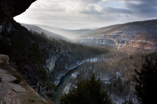 A scenic view of a river winding through a snowy valley, surrounded by misty mountains under a cloudy sky.