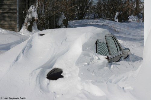 A car almost completely buried in deep snow, with only the roof and windows visible. Surrounding landscape is snowy.