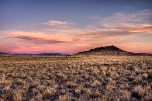 A vast, open landscape at sunset, featuring golden grass and a distant hill under a colorful sky.
