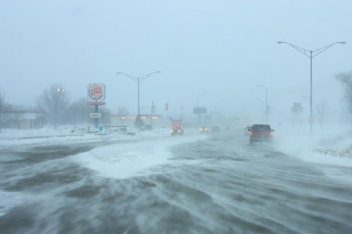 A snowy road scene with low visibility, cars navigating through a winter storm, and a Burger King sign in the background.