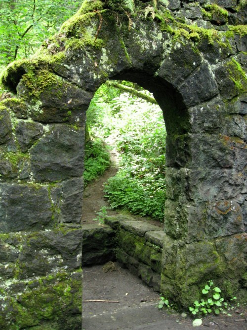A moss-covered stone archway leads to a lush, green forest path.