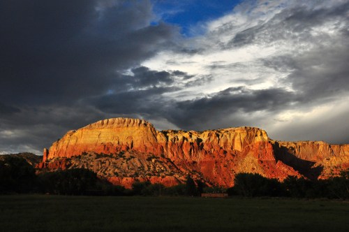A dramatic sunset illuminates a rugged mountain landscape, casting warm hues on the rocky cliffs under a cloudy sky.