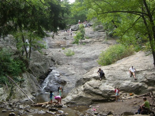 A rocky landscape with a small waterfall, people relaxing and playing near the water, surrounded by lush greenery.