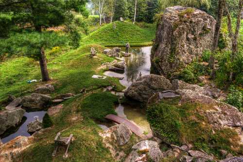 A serene landscape featuring a pond, rocks, and a person tending to the garden amidst lush greenery.