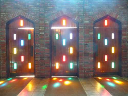 Three wooden doors with colorful glass panels, set against a brick wall, casting vibrant light patterns on the floor.