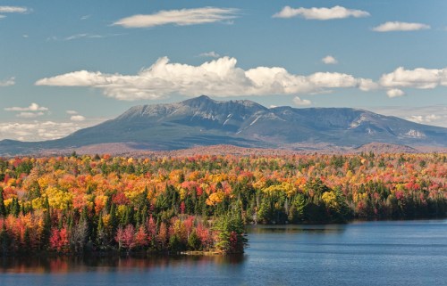 A vibrant autumn landscape featuring colorful trees, a serene lake, and a mountain under a blue sky with fluffy clouds.