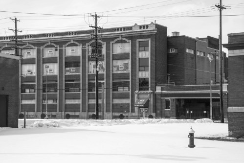 A large brick building with multiple windows, surrounded by snow and utility poles, under a cloudy sky.