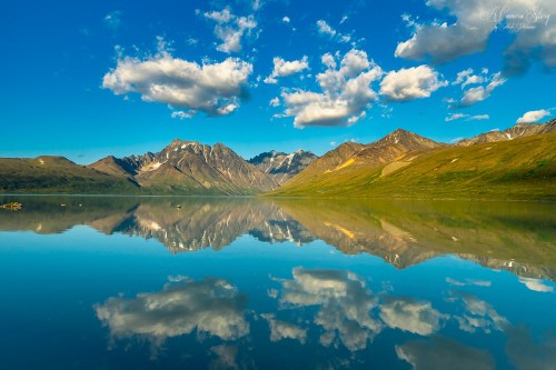 A serene mountain landscape reflected in calm water, with blue skies and fluffy clouds above.