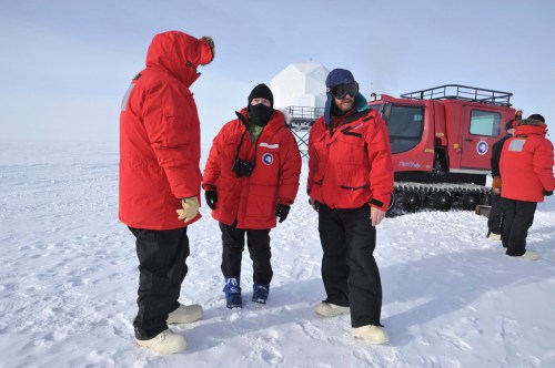 Three people in red winter gear stand on snow, with a vehicle in the background against a clear sky.