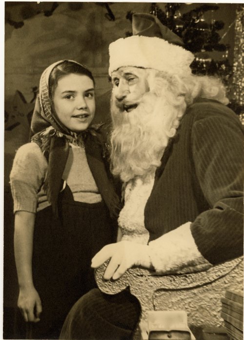 A young girl in a scarf smiles next to Santa Claus, who is seated and wearing a traditional red suit and hat.