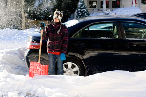 A child in a winter hat and gloves stands with a shovel beside a car, surrounded by snow.
