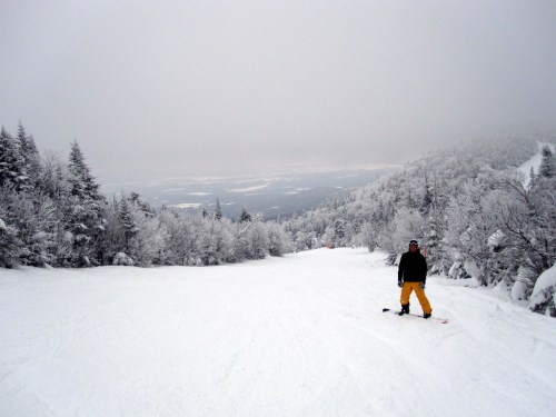 A snowboarder in yellow pants stands on a snowy slope surrounded by frosted trees under a cloudy sky.