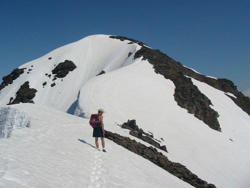 A hiker in shorts and a backpack walks on a snowy mountain trail under a clear blue sky.