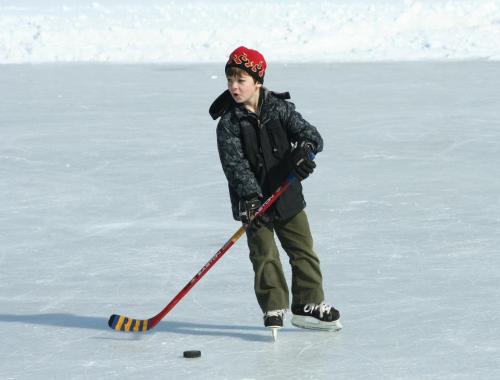 A young boy in a black jacket and red hat skates on ice, preparing to hit a hockey puck with his stick.