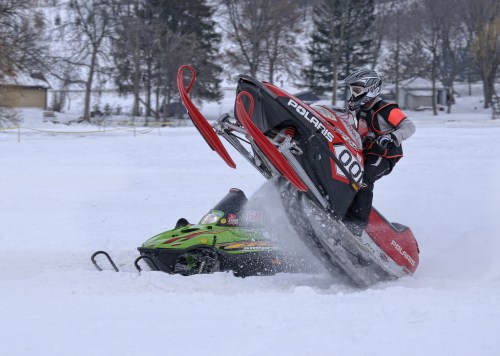 A snowmobiler performs a stunt, lifting the front of the sled off the ground in a snowy landscape.