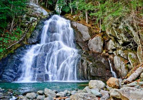 A serene waterfall cascading over rocks into a clear pool, surrounded by lush greenery and rugged terrain.