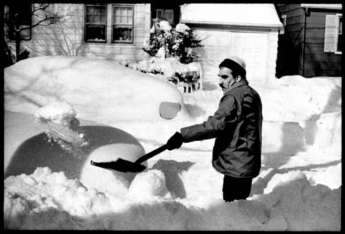 A man shovels snow from a car in a snowy residential area, with snow-covered houses in the background.