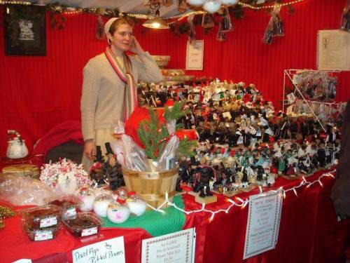 A vendor stands at a festive market stall filled with holiday decorations and crafts, surrounded by red and green decor.