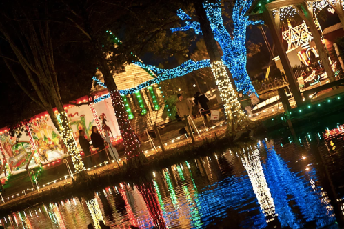 Colorful holiday lights reflect on a calm water surface, with people enjoying the festive atmosphere in a park.