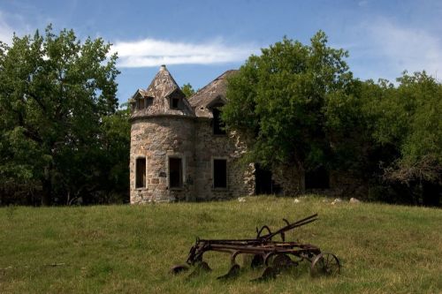 An abandoned stone house with a turret, surrounded by trees and an old plow in a grassy field under a blue sky.
