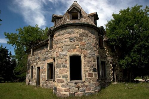 Abandoned stone house with a turret, surrounded by grass and trees, under a partly cloudy sky.