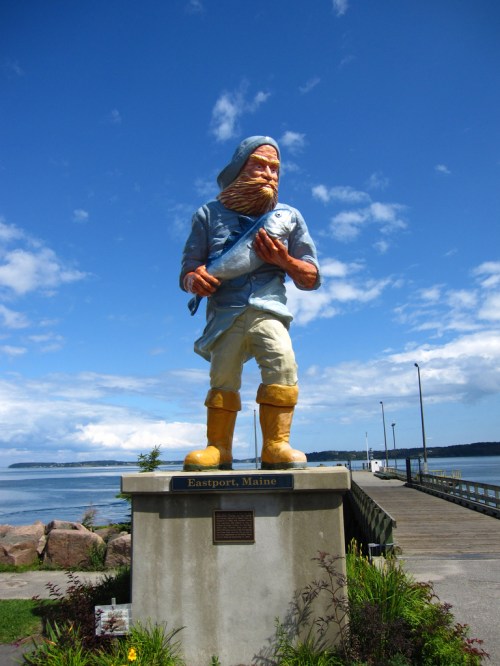 A large statue of a fisherman stands in Eastport, Maine, with a scenic waterfront and blue sky in the background.