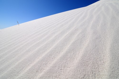 A smooth, white sand dune under a clear blue sky, showcasing gentle ripples in the sand.