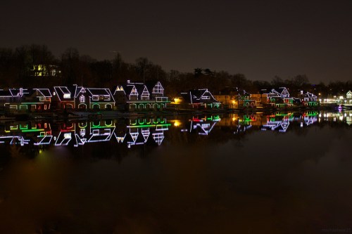 Colorful holiday lights adorn houses along a river, reflecting beautifully on the water at night.