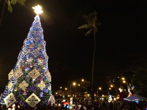 A brightly lit Christmas tree adorned with colorful decorations, set against a night sky with festive crowds below.