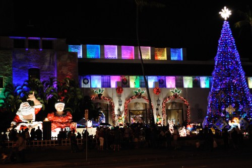 Colorful Christmas lights illuminate a festive scene with a large tree, Santa figures, and a crowd enjoying the decorations.