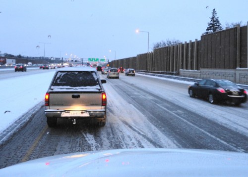 A snowy highway scene with vehicles driving cautiously on a slick road, surrounded by snow-covered barriers.