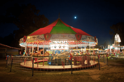 A brightly lit carousel at night, surrounded by festive lights and a full moon in the sky.