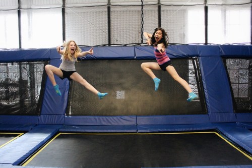 Two girls joyfully jumping on a trampoline, mid-air, with big smiles and arms outstretched.