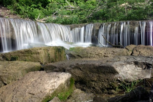 A serene waterfall cascades over rocks, surrounded by lush greenery and a tranquil natural setting.