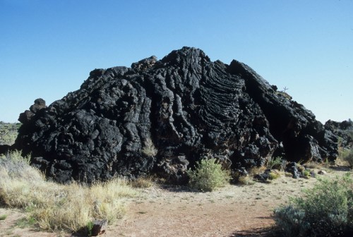 A large, dark volcanic rock formation surrounded by sparse vegetation under a clear blue sky.