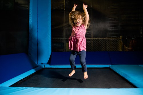A joyful child with curly hair jumps on a trampoline, arms raised in excitement, against a dark background.