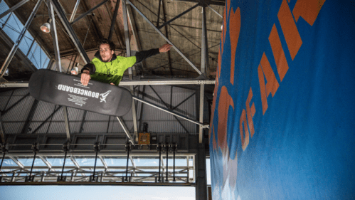 A person in a green shirt performs a trick on a snowboard, soaring above a blue wall with "Out of Air" written on it.