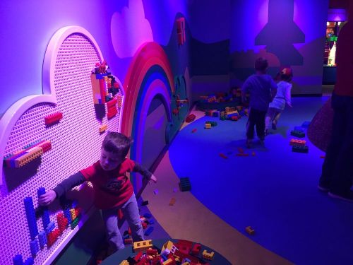 Children play in a colorful room with LEGO bricks, building structures against a wall and exploring the space.