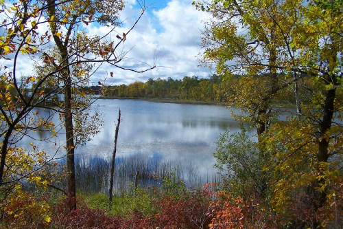 A serene lake surrounded by trees with autumn foliage under a partly cloudy sky. Reflections shimmer on the water's surface.